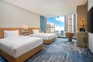 A hotel room with two beds, a desk, a chair, and a view of tall buildings.