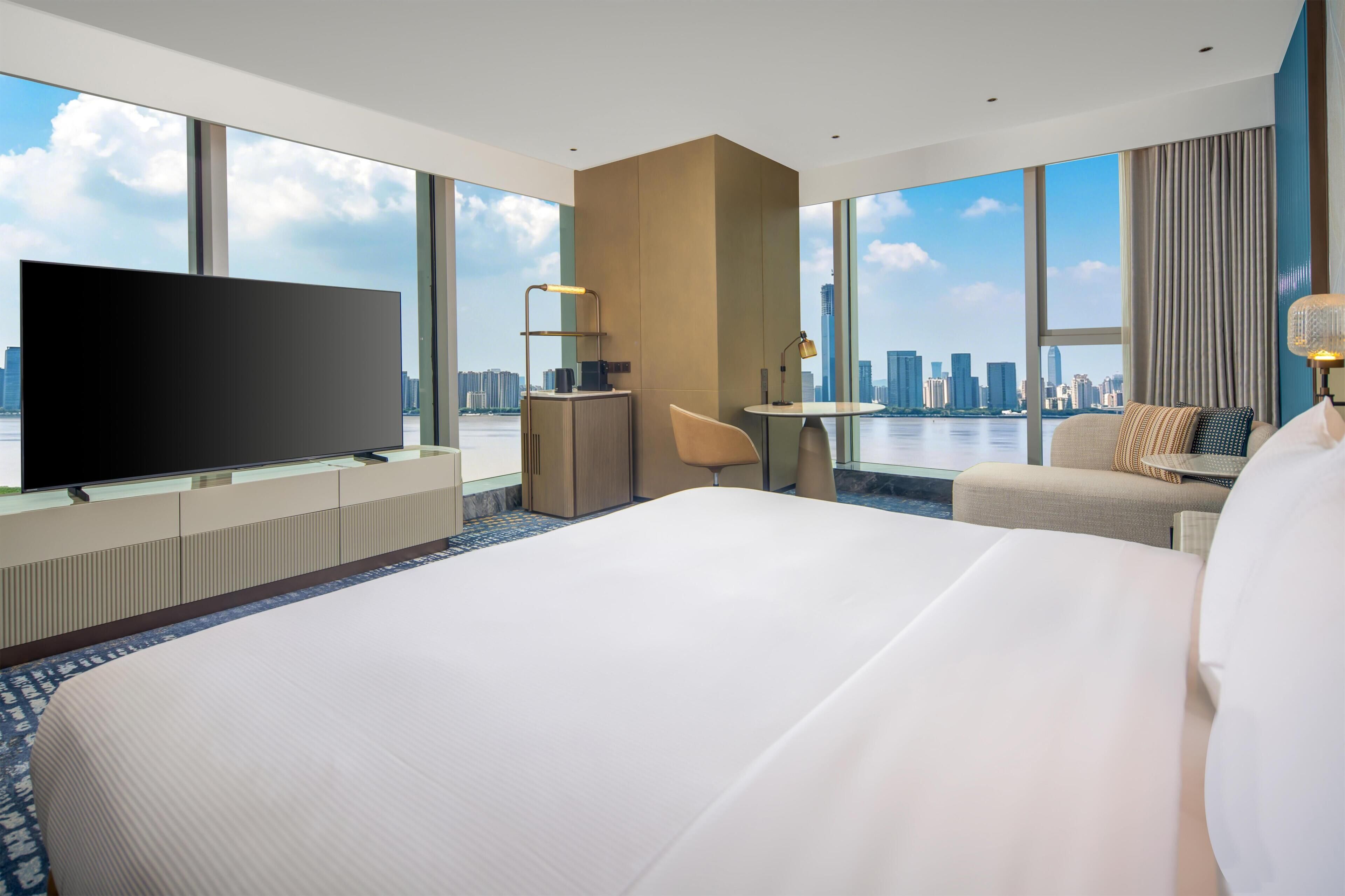 A hotel room with a large bed, a TV, a desk, and a view of the city skyline.