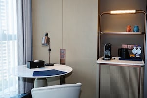Quarto de hotel com uma mesa redonda, uma cadeira, um abajur, uma cafeteira e uma estante com chávenas.