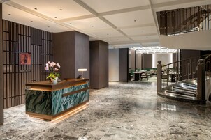 Um hall de hotel com balcão de recepção, piso de mármore, escada e área de estar.