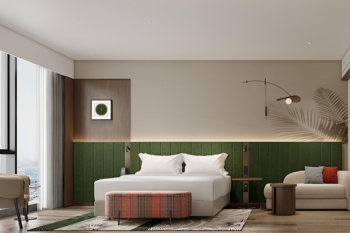 Habitación de hotel moderna con una cama grande, una zona de estar, vistas a la ciudad y una pared de acento verde.