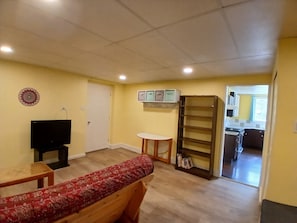 Ein Wohnzimmer mit einer Couch, einem Fernseher, einem kleinen Tisch, einem Bücherregal und einer Tür, die in eine Küche führt.