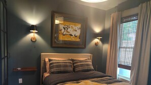 Un dormitorio con cama, dos lámparas de pared, una obra de arte enmarco, una ventana con cortinas y una mesita de noche con lámpara.