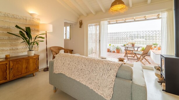 A cozy living room with a sofa, a wooden cabinet, a potted plant, a hanging light fixture, and a view of the patio area. - Casinha do Burro – Casas do Poente: Cozy Cottage with Private Patio (Rogil)