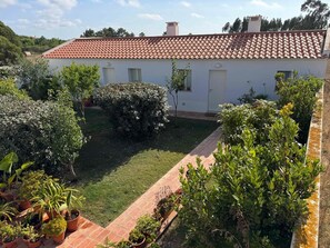 A white building with a red-tiled roof, surrounded by a well-maintained garden with various potted plants and a grassy lawn. - Casinha do Burro – Casas do Poente: Cozy Cottage with Private Patio (Rogil)
