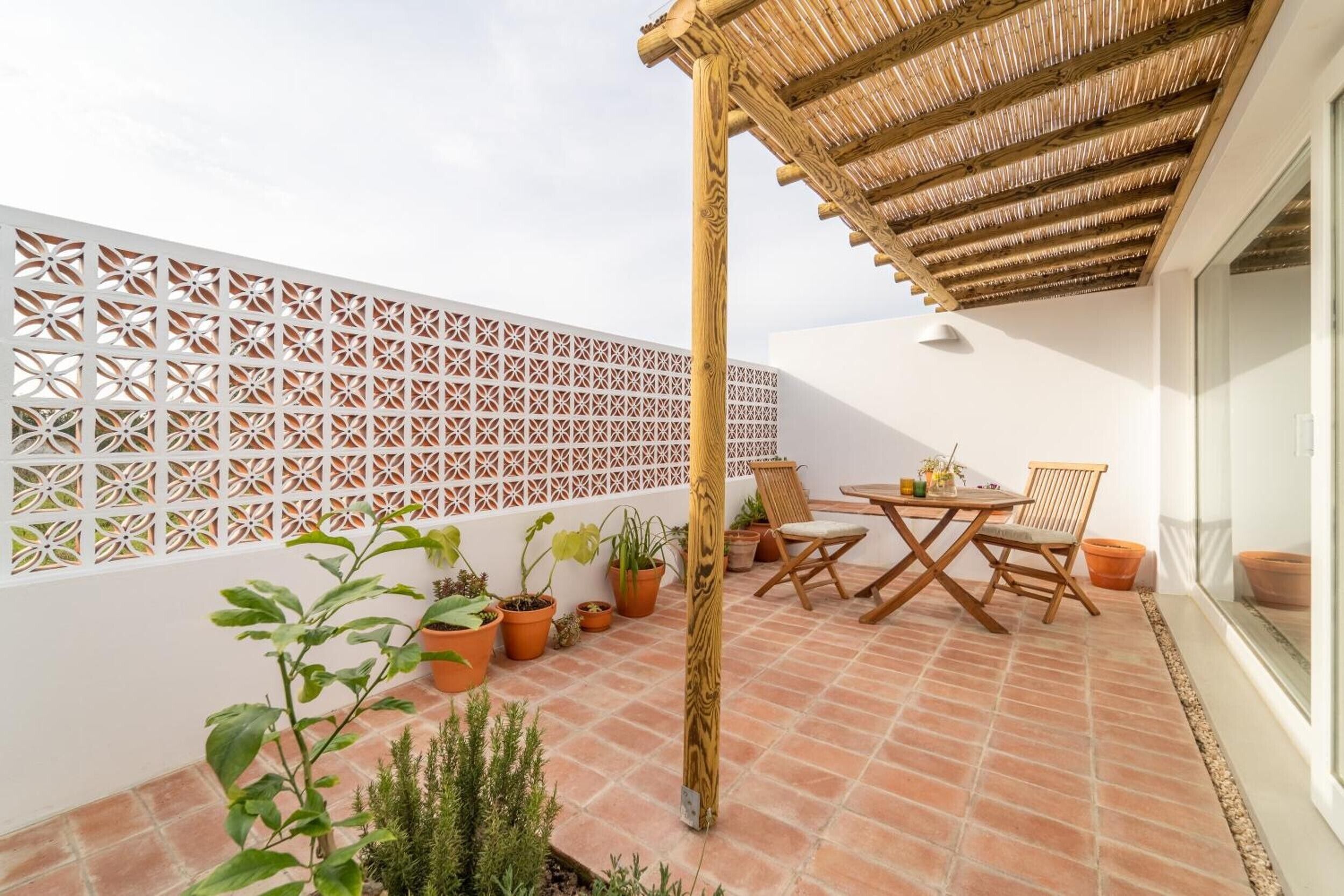 An outdoor patio with a table and chairs, potted plants, a wooden pergola, and a decorative white lattice wall.
