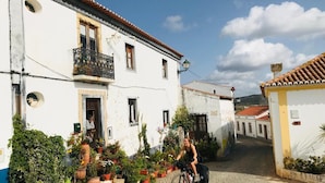 A narrow cobblestone street lined with white buildings, a cyclist, and potted plants. - Casinha do Burro – Casas do Poente: Cozy Cottage with Private Patio (Rogil)