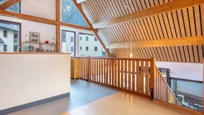 A spacious attic room with a sloped ceiling, wooden beams, a view through large windows, and a wooden railing along the floor. - Mountain Lodging With Private Balcony (Saint-Colomban-des-Villards)