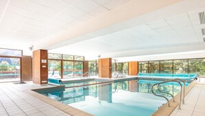 Una grande piscina interna con pareti di vetro trasparente, pavimento piastrellato e vista sul verde circostante.