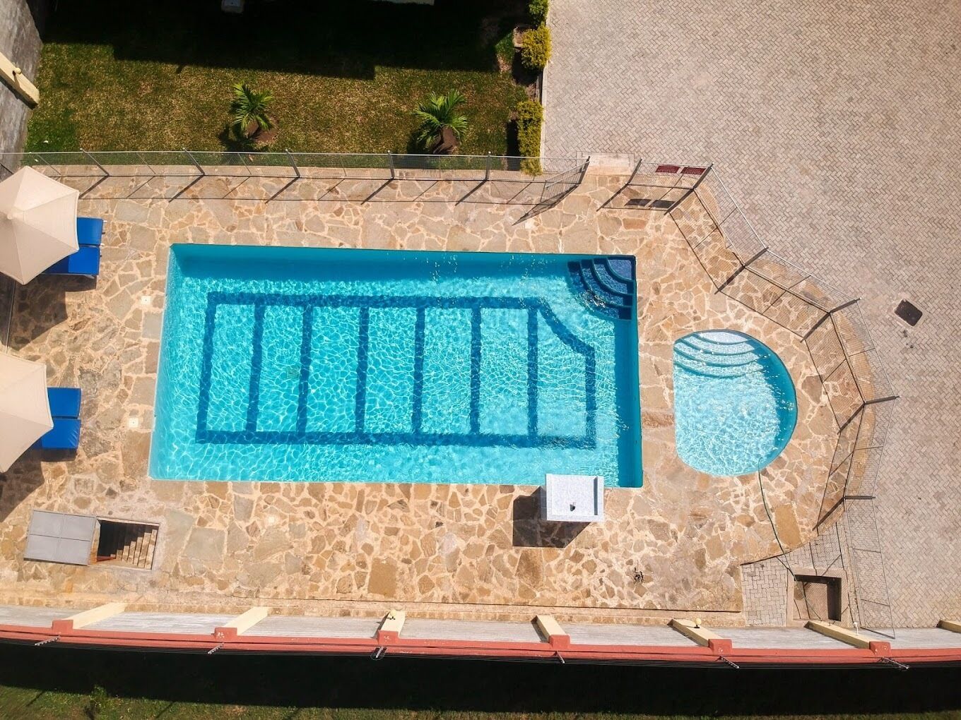 An aerial view of a swimming pool with a stone deck, a small circular pool, and a grassy area with palm trees.