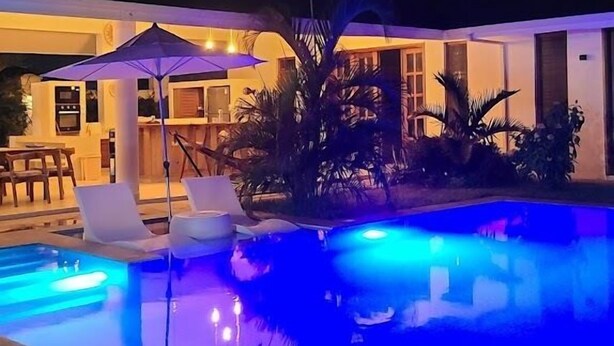 Una piscina con iluminación azul, un área de patio con sillas y un edificio con grandes ventanales al fondo.