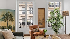 A living room with a sofa, armchair, coffee table, and a large window with a view of buildings. - Gourmandise, 2BR/1BA, 4 people (Paris)