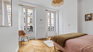 A bedroom with a bed, a chair, a radiator, and a window with a view of buildings. - Gourmandise, 2BR/1BA, 4 people (Paris)