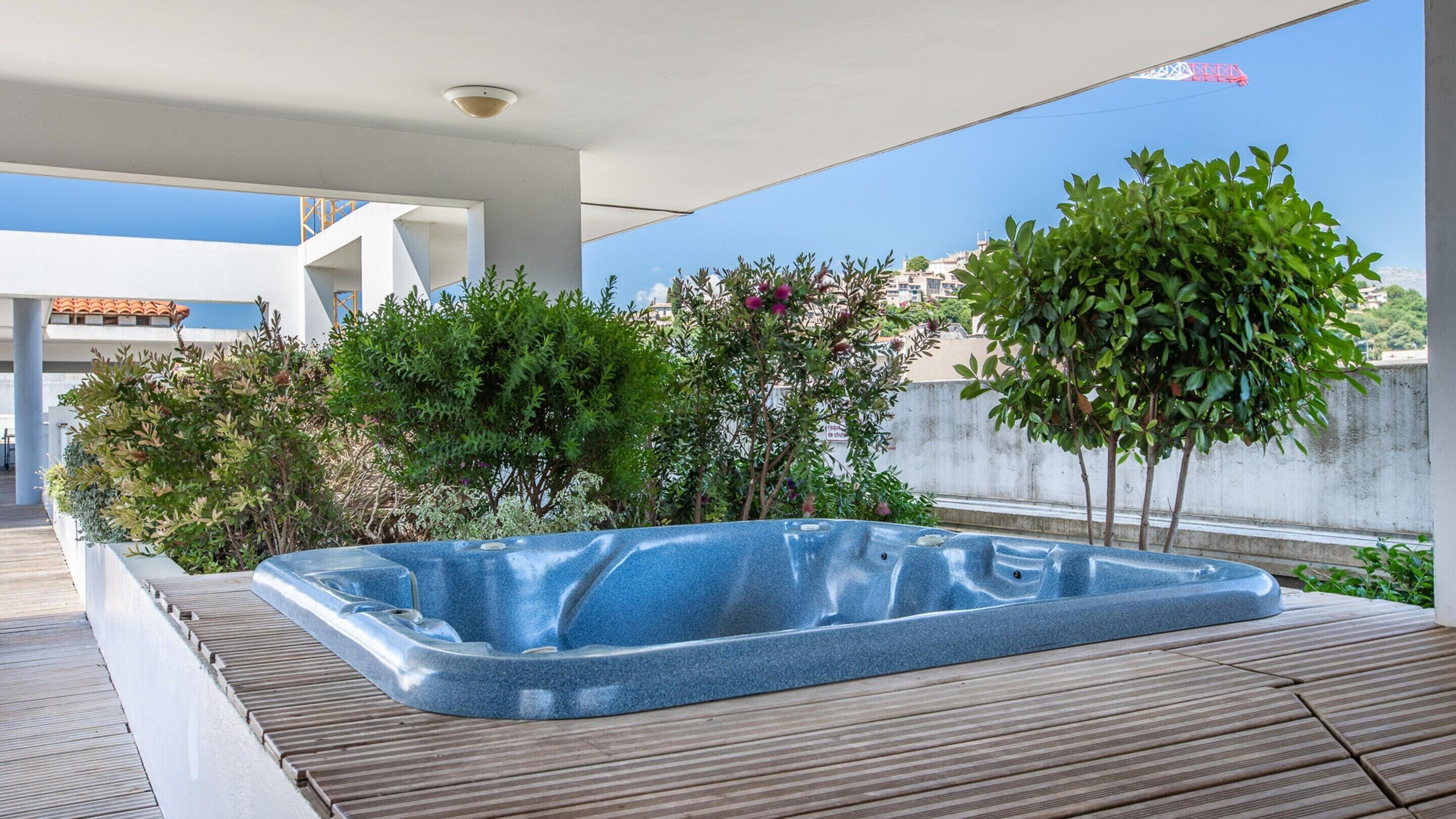 Un jacuzzi en una terraza de madera rodeado de follaje verde y un cielo despejado.