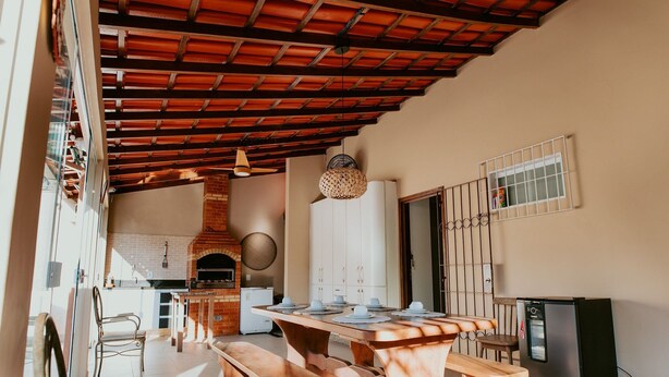 An open-plan kitchen with a wooden table, benches, a refrigerator, and a brick oven. - Cozy House with Gourmet Area and Pool (Vila Velha)