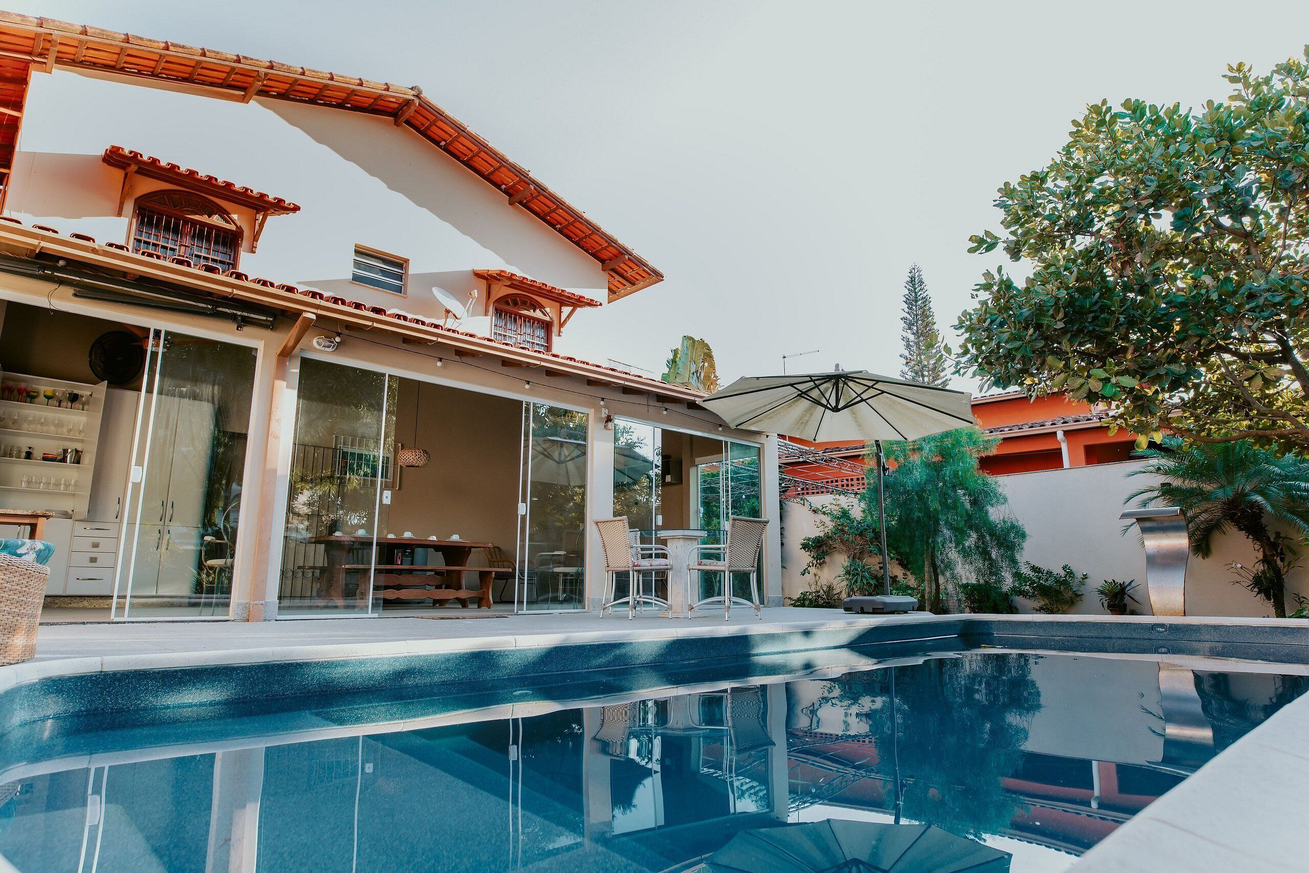 Une piscine entourée de parois en verre, un patio couvert et un bâtiment doté d’un toit en tuiles et de grandes fenêtres.