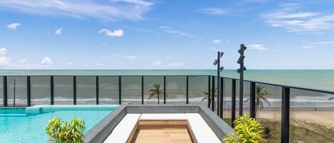 A rooftop pool with a wooden deck, surrounded by a glass balustrade, overlooking a beach and ocean.