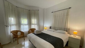 Ett hotellrum med en säng, två stolar, ett nattduksbord med en lampa och tunna, genomskinliga gardiner.