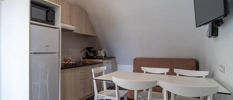 Une cuisine compacte équipée d’un réfrigérateur, d’un four à micro-ondes et d’une machine à café. On y trouve deux chaises blanches et une petite table. Un téléviseur à écran plat est fixé au mur.