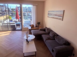 Ein modernes Wohnzimmer mit einem grauen Sofa, einem kleinen weißen Tisch mit einer Pflanze und Blick auf eine Marina durch große Glasüren.