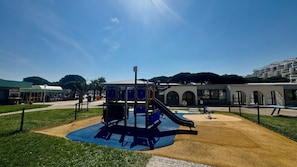 A playground with a slide and climbing structure, surrounded by a fenced area with a building in the background. - Le Port Carnon, Parenthèse Marine (Carnon-Plage)