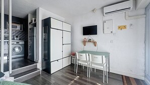 Een compacte kamer met kitchenette, een eethoek met tafel en stoelen, een wandgemonteerde televisie en een airconditioning.