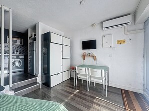 A compact room with a kitchenette, a dining area with a table and chairs, a mounted TV, and an air conditioning unit. - Le Port Carnon, Parenthèse Marine (Carnon-Plage)