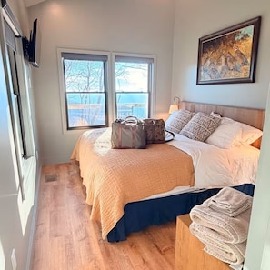 A bedroom with a large bed, two windows, a painting, and a wooden floor.