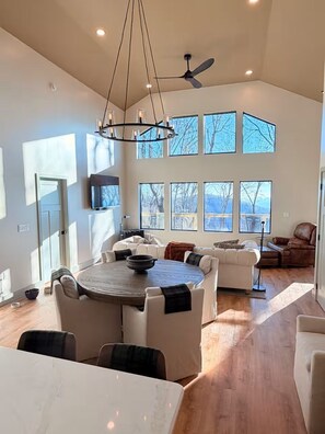 A spacious living area with a high ceiling, large windows, a dining table, and a ceiling fan.