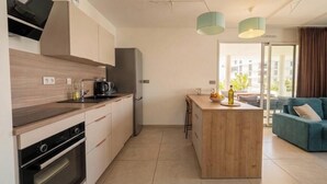 A modern kitchen with a central island, a dining area with a blue sofa, and a view of buildings through a glass door. - L'apogée, centre et terrasse (Montpellier)