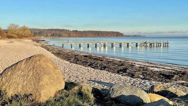 Een strand met een houten pier die uitsteekt in kalm water, omgeven door rotsen en schaarse begroeiing.