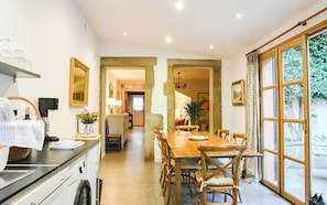 A dining area with a wooden table and chairs, a kitchen area with a counter and appliances, and a view through a large window. - Charming riverside house in medieval village (Villeneuve-Minervois)