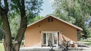 A small wooden cabin with a sliding glass door, a patio with metal chairs, and a large tree to the left.