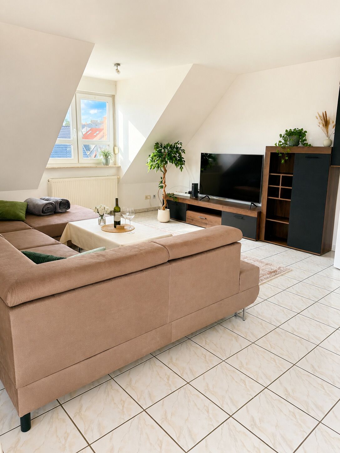 A modern living room with a sectional sofa, a flat-screen TV, a potted plant, and a dining table with a wine bottle.