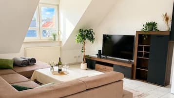 A modern living room with a sectional sofa, a flat-screen TV, a potted plant, and a dining table with a wine bottle.