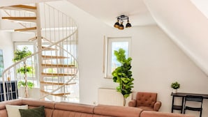 A modern living room with a sectional sofa, a dining table with a bottle of wine and grapes, a chair, and a staircase. - Velmora Boutique Suites Rheingau - Balcony & Grill (Oestrich-Winkel)