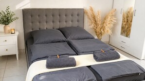 A modern bedroom with a tufted headboard, a large bed with grey bedding, a white nightstand, a mirror, and a fluffy rug. - Velmora Boutique Suites Rheingau - Balcony & Grill (Oestrich-Winkel)