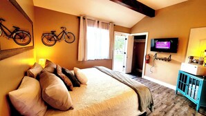 Une chambre à coucher avec un lit, des oreillers, une télévision, un vélo et une porte qui donne sur une autre pièce.