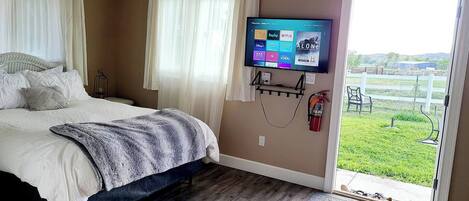 A bedroom with a bed, curtains, a mounted TV displaying streaming services, a fire extinguisher, and a view of a grassy area through a door.