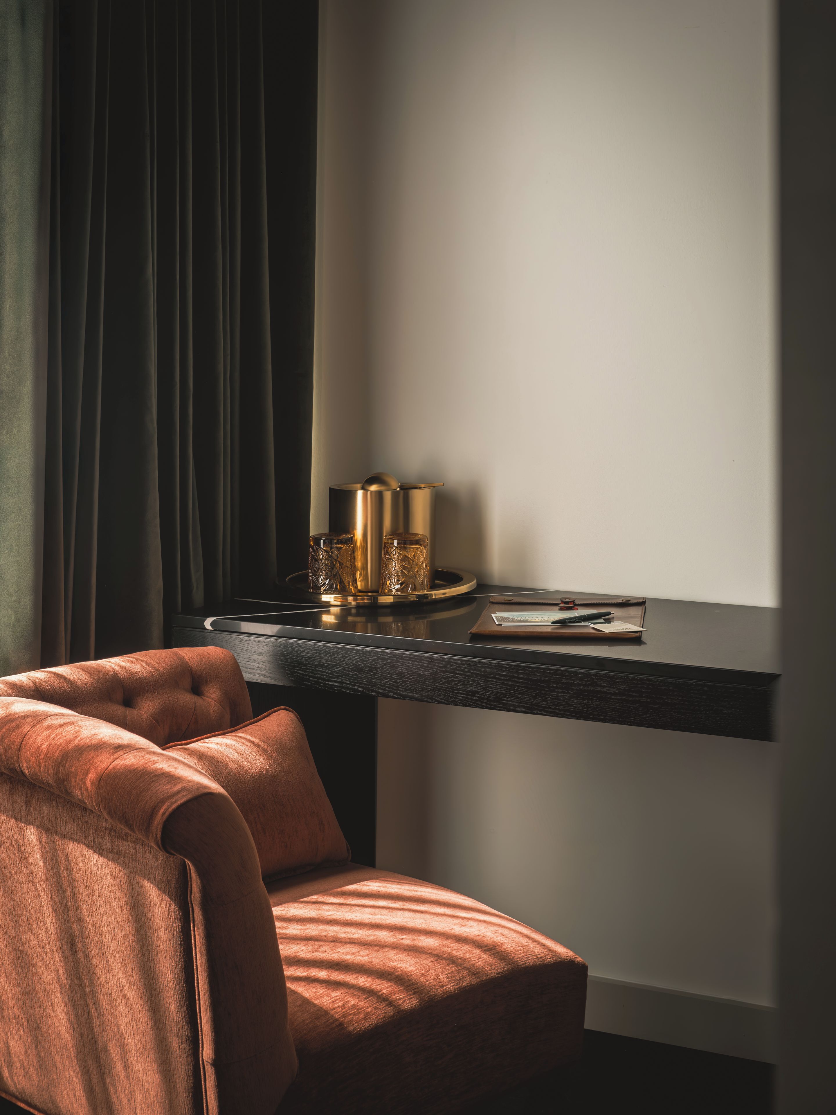 A room with a dark curtain, a tufted armchair, a desk with a tray, and a lamp.