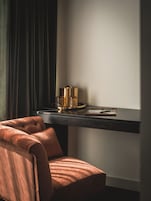 Una habitación con una cortina oscura, un sillón capitoné, un escritorio con una bandeja y una lámpara.