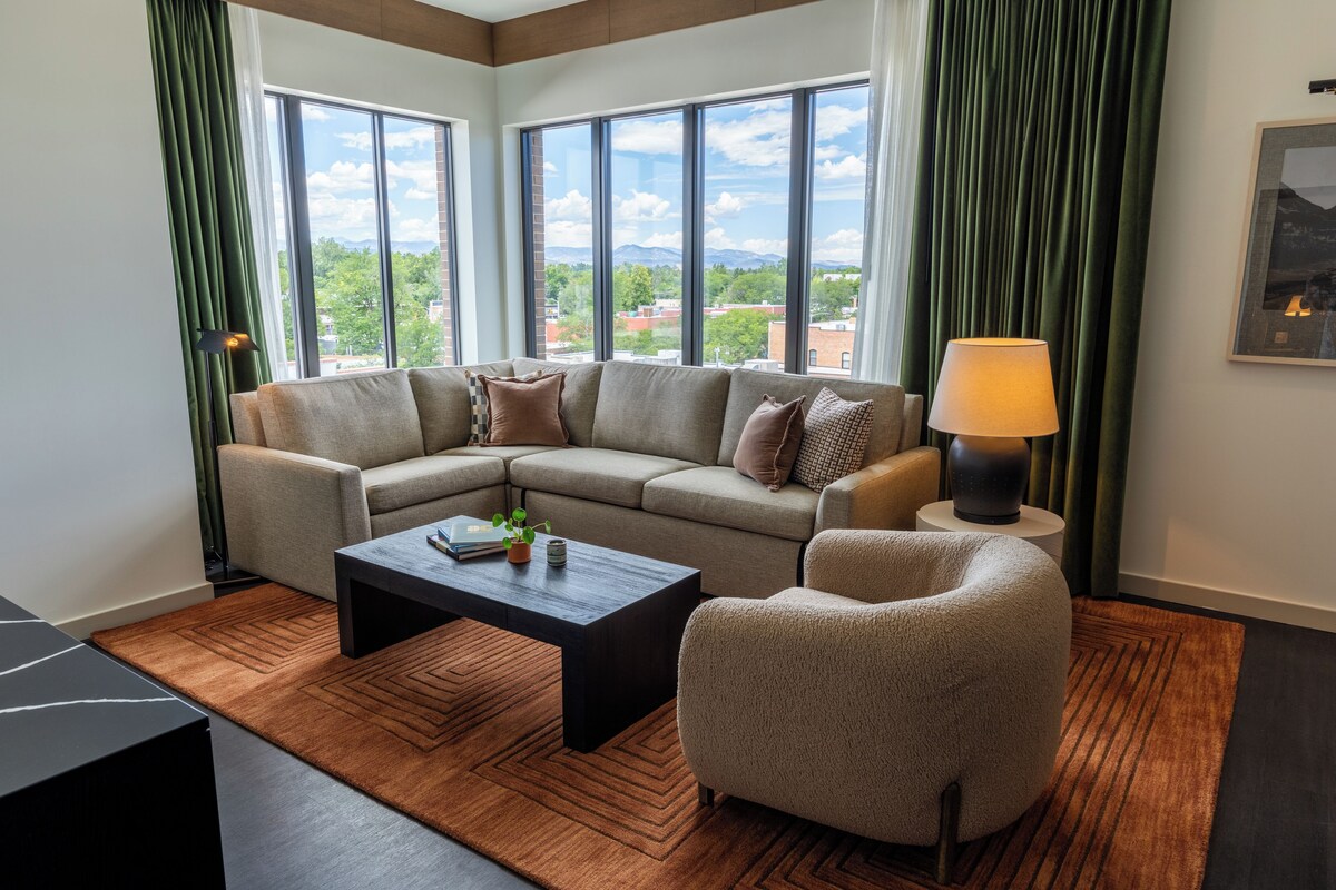 A modern living room with a sectional sofa, a coffee table, a side chair, and a lamp. Large windows provide a view of the cityscape.