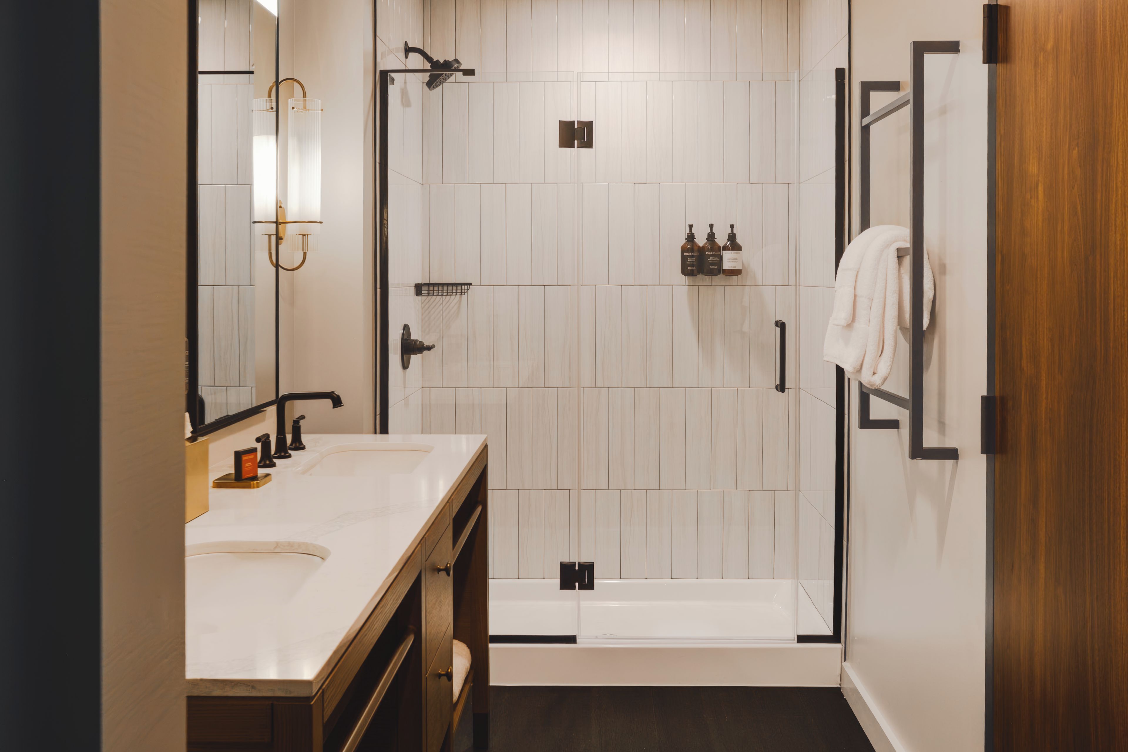 A modern bathroom with a double sink vanity, a glass shower enclosure, and a wooden door.