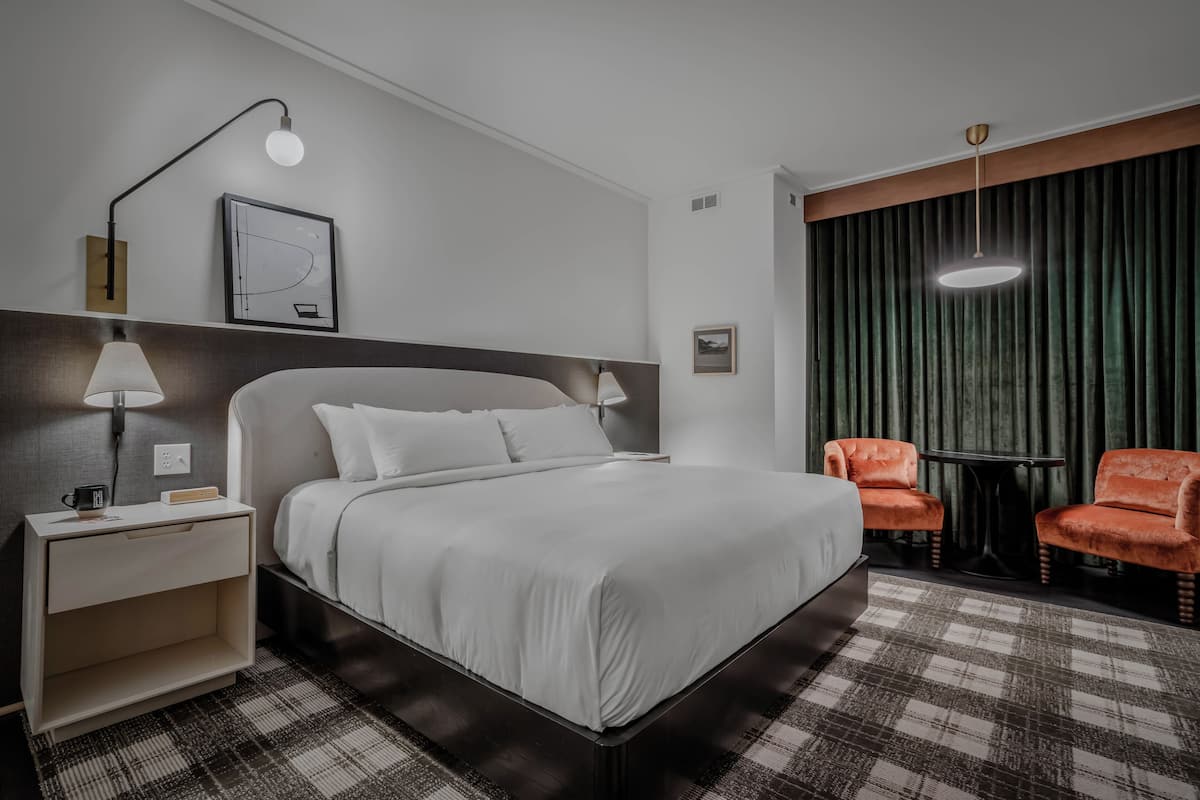 A hotel room with a bed, a nightstand, a lamp, a framed picture, a window with curtains, and two chairs.