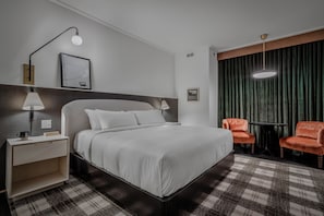 A hotel room with a bed, a nightstand, a lamp, a framed picture, a window with curtains, and two chairs. - Hotel Longmont (Longmont)
