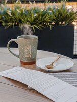 Una mesa con una taza de cerámica, un plato con una cuchara y un menú.