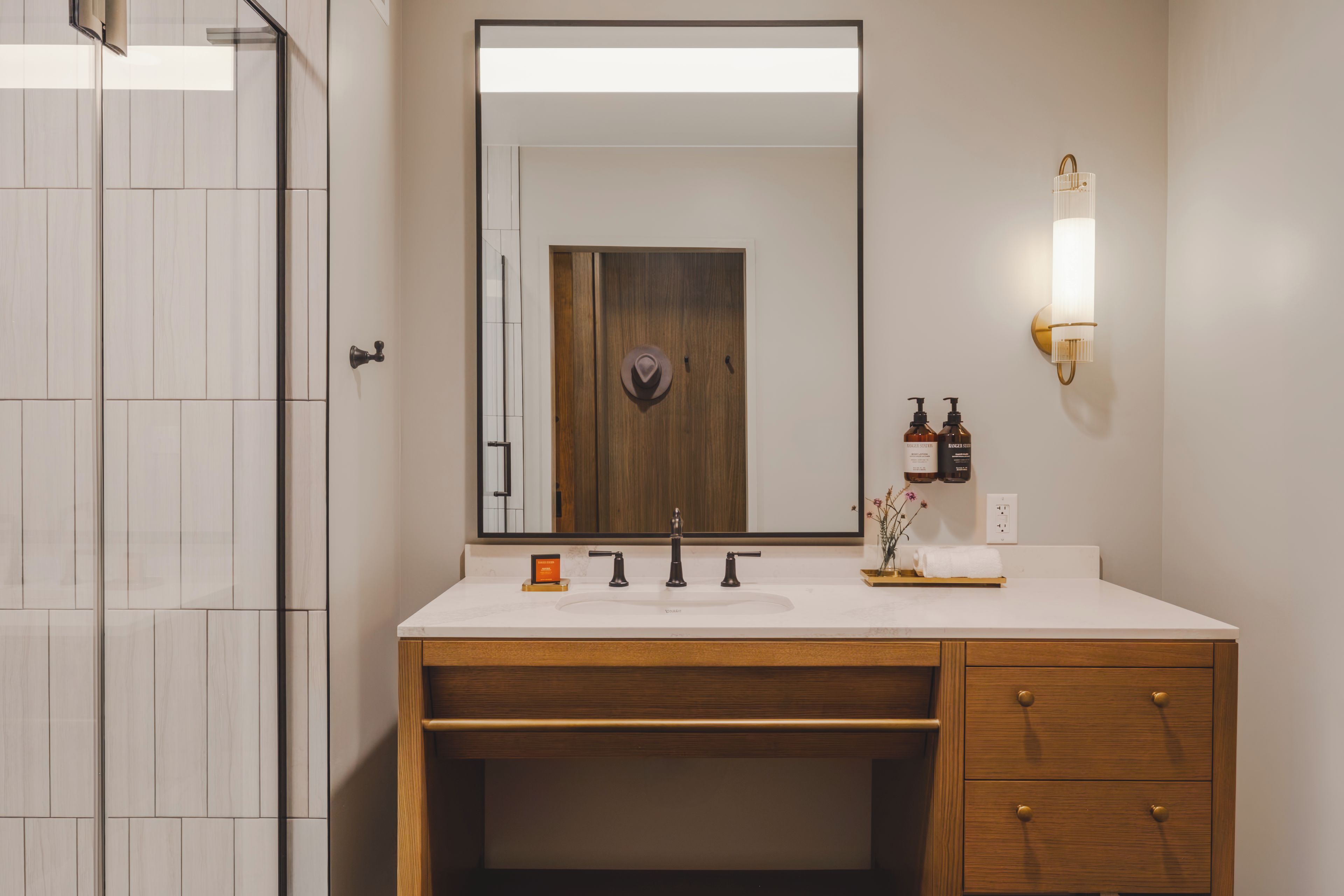 Un baño con tocador de madera, un espejo grande, una lámpara en la pared y una zona de ducha.