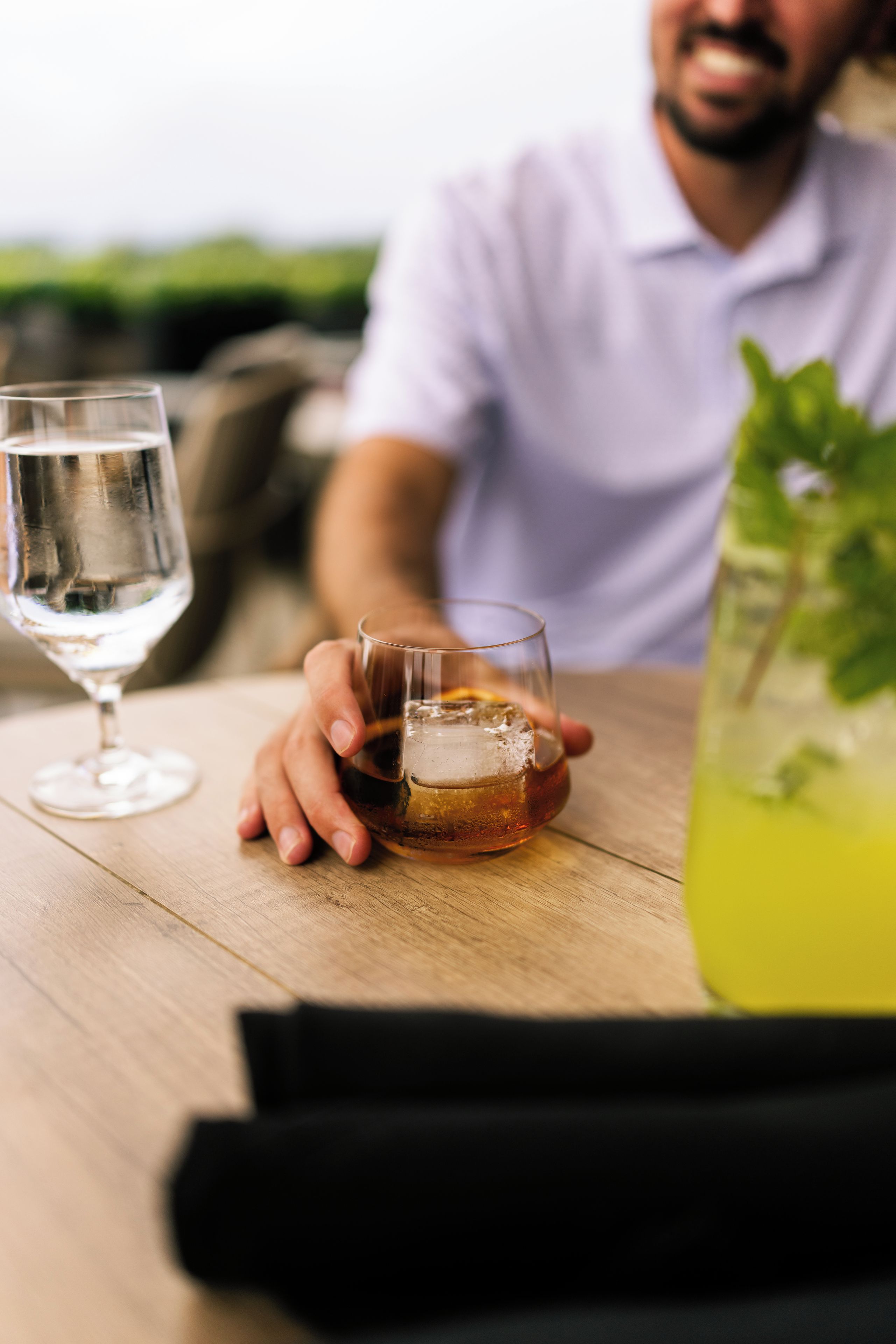 Une personne tenant un verre contenant un liquide ambré, avec un verre transparent et un verre d’eau posés sur la table.