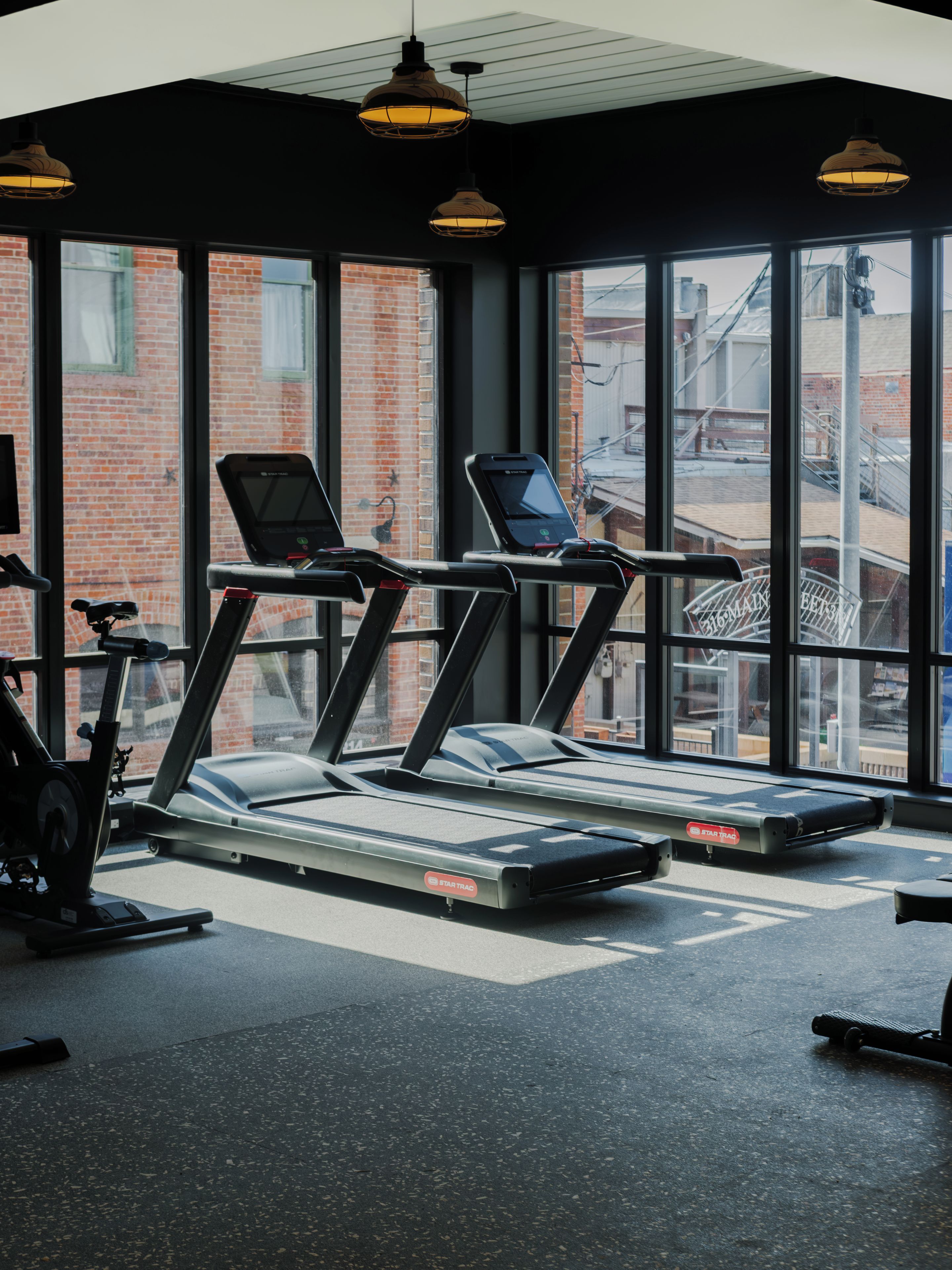 Un gimnasio con cintas de correr y bicicletas estáticas, ventanas amplias y una pared de ladrillo en el exterior.