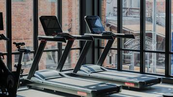 Un gimnasio con cintas de correr y bicicletas estáticas, ventanas amplias y una pared de ladrillo en el exterior.